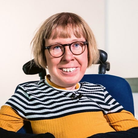 Anne Gersdorff, eine Frau mit kurzen blonden Haaren, die in einem Rollstuhl sitzt und lächelt. Sie trägt einen gelb, weiß, schwarzen Pulli und eine schwarze Brille.